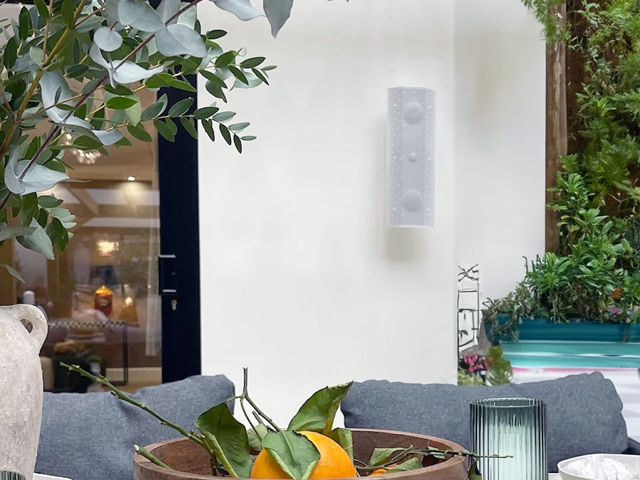 A white Lithe Audio outdoor speaker on a wall beside a pool and planting