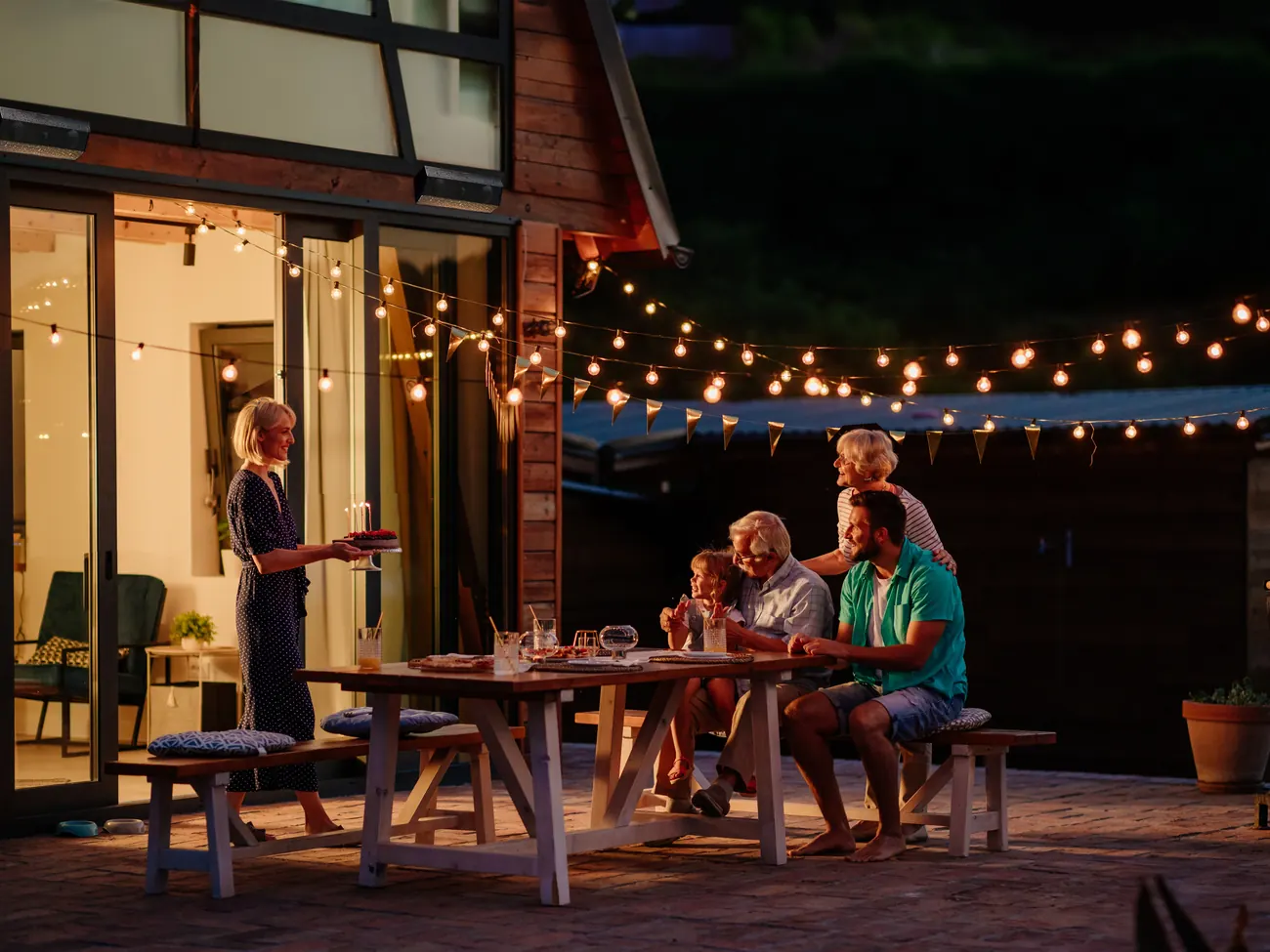 Friends eating at a long table on a terrace at night under festoon lights